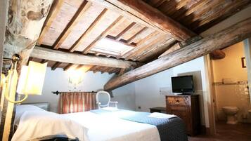 Una habitación rústica con techo de madera, una cama con cubrecama blanco, un escritorio con teléfono y un televisor sobre un mueble de madera.