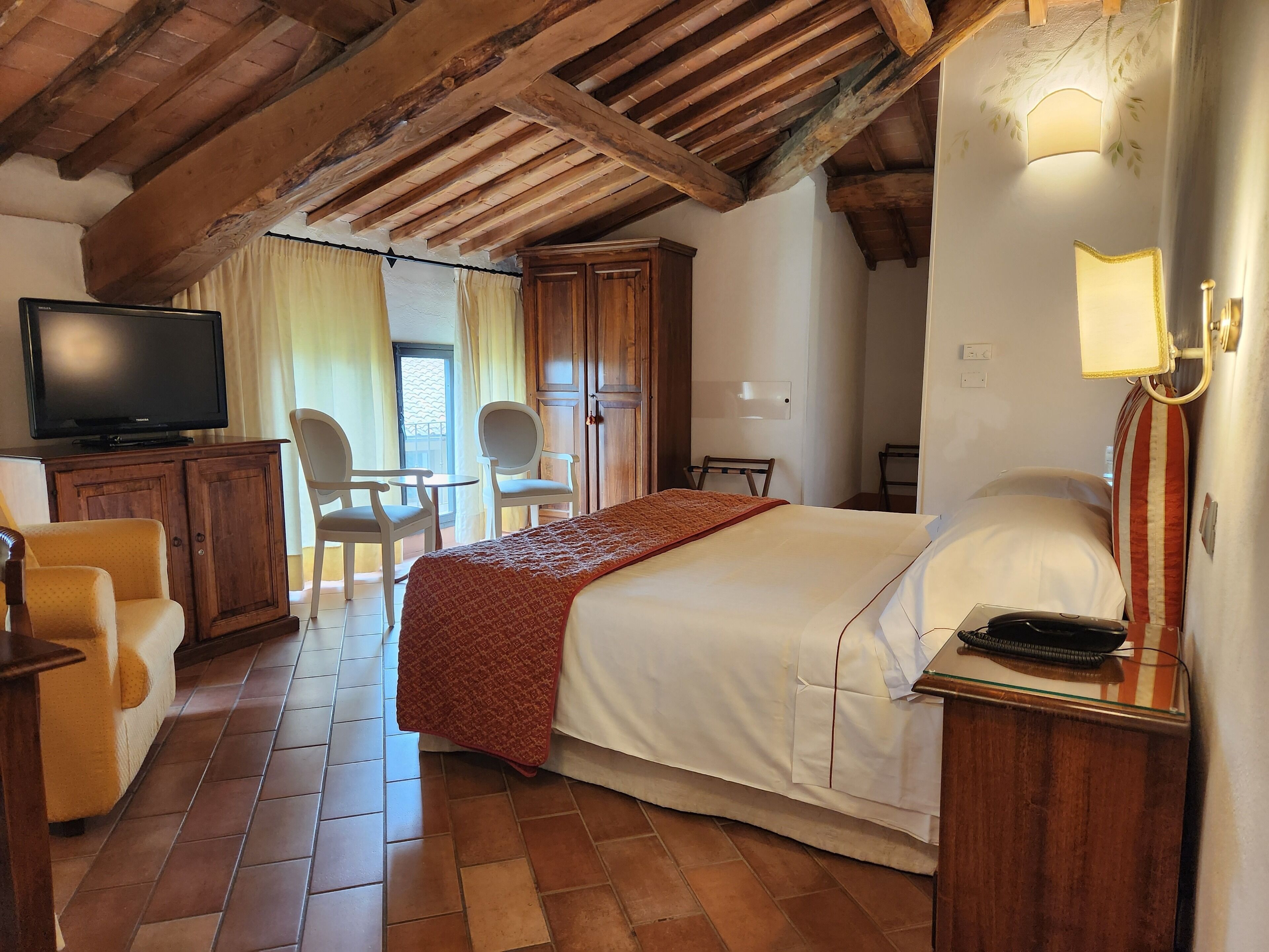 A hotel room with a bed, a television, a desk with a lamp, a chair, and a wardrobe.