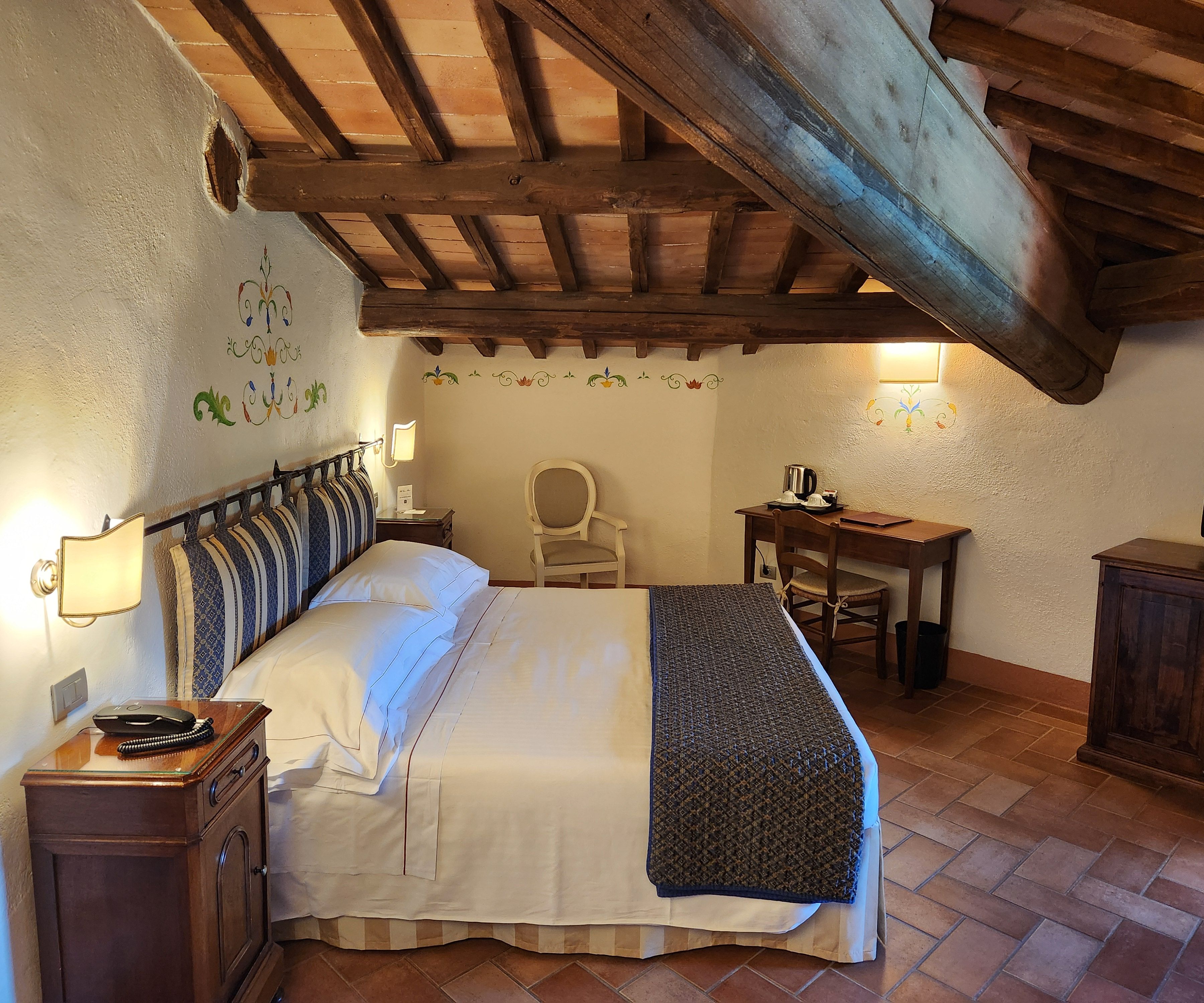 A bedroom with a large bed, a desk, a chair, and a small table with a lamp.