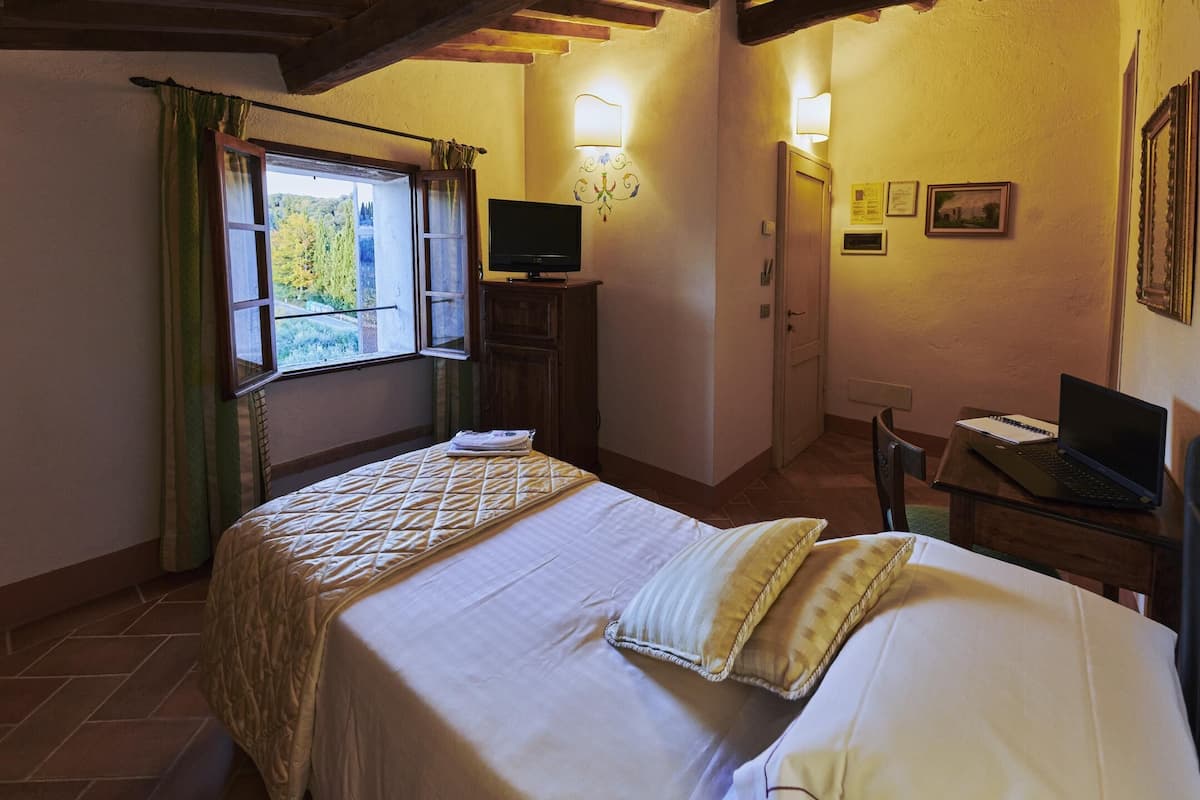 Un dormitorio con una cama, televisor, escritorio con una computadora portátil y ventana con cortinas.