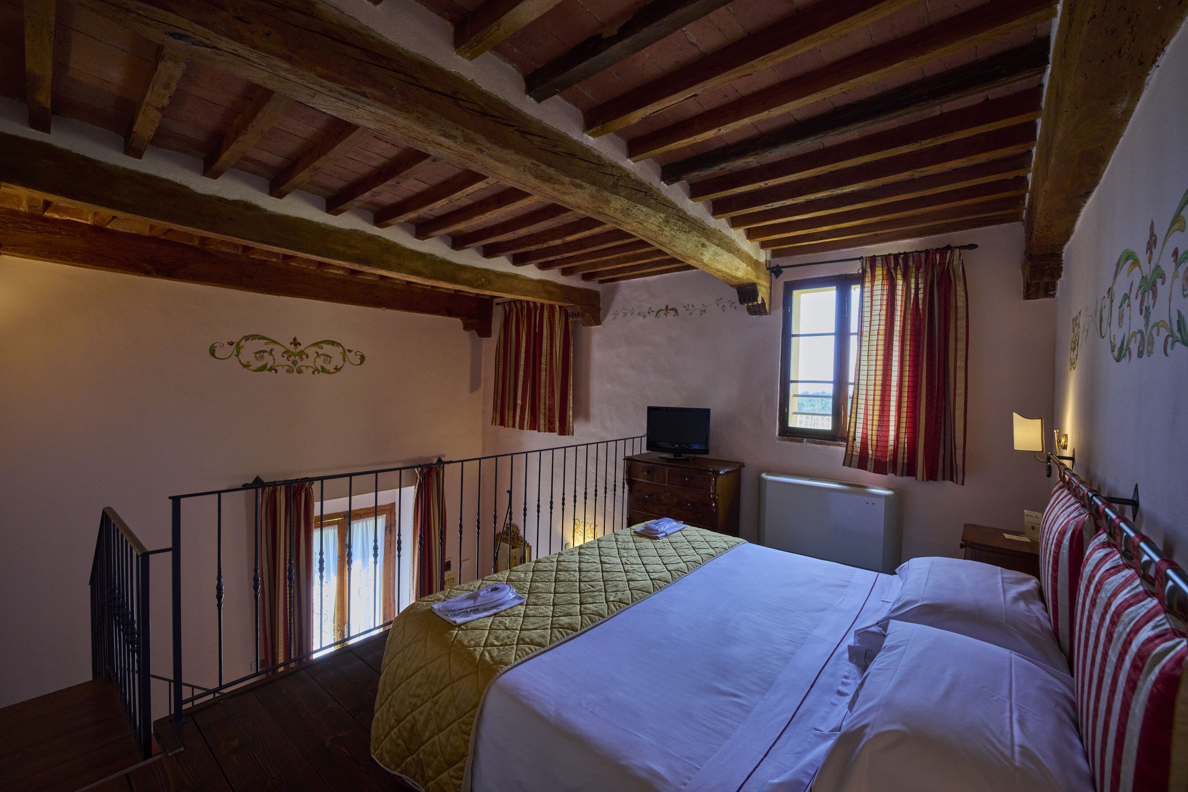A bedroom with a bed, wooden beams, a TV, a window with curtains, and a radiator.