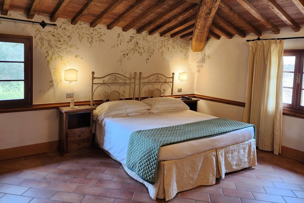 Un dormitorio con techo de madera, una cama con manta verde, dos mesitas de noche y una ventana con cortinas.