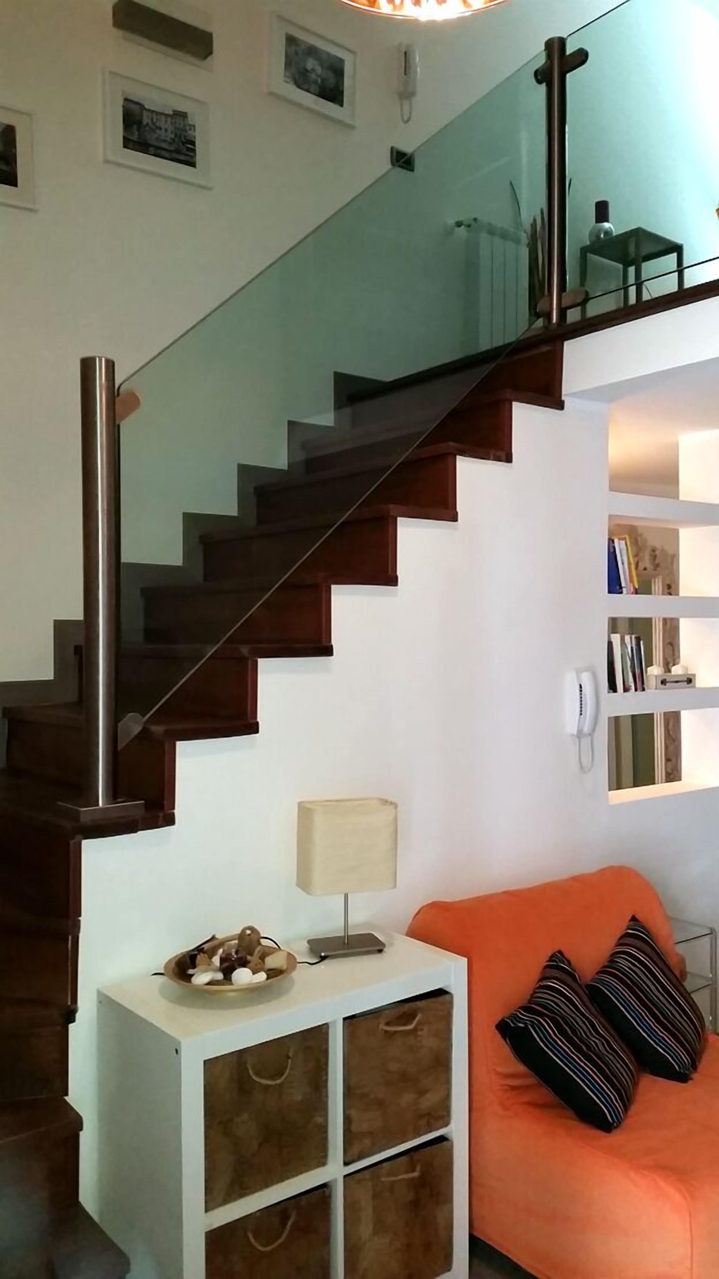 Una escalera moderna con barandillas de vidrio, una mesita auxiliar blanca con lámpara, y un sofá naranja con cojines a rayas.