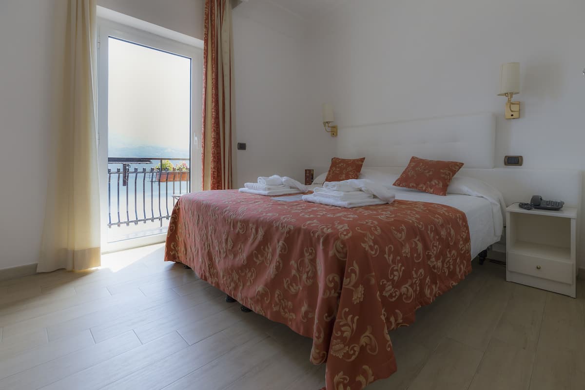 Habitación de hotel con una cama grande, una mesita de noche, una lámpara de pared y vistas al balcón.