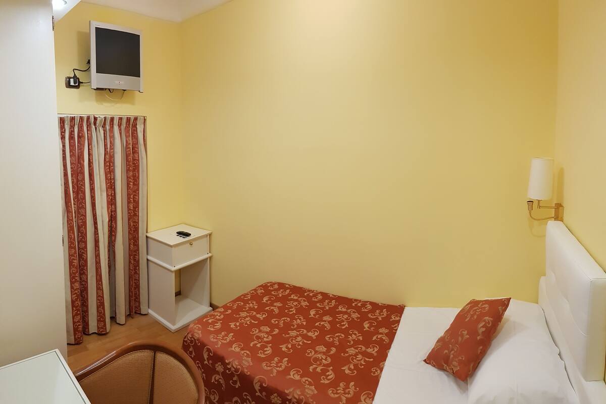 Una habitación pequeña con paredes amarillas, una cama individual, un escritorio, un televisor y una lámpara.