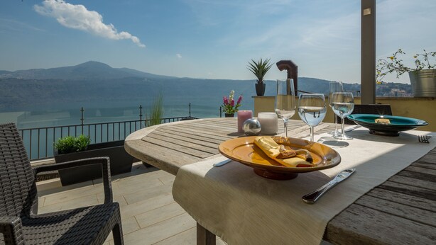 Una mesa dispuesta con comida, vasos y una vela en una terraza con vistas a un lago y montañas.