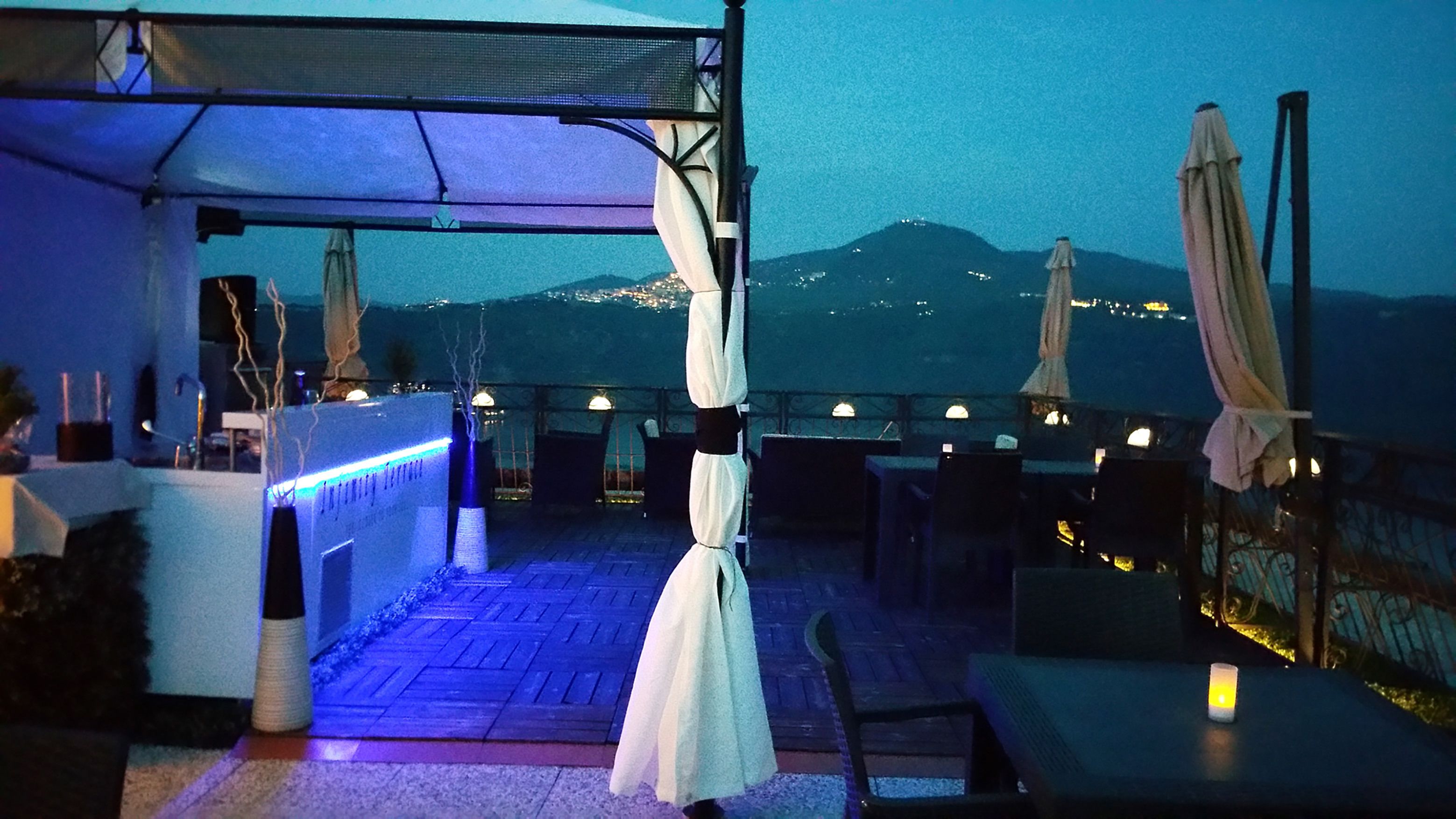 Una terraza al aire libre con mesas y sillas, sombrillas y vistas a la montaña al atardecer.