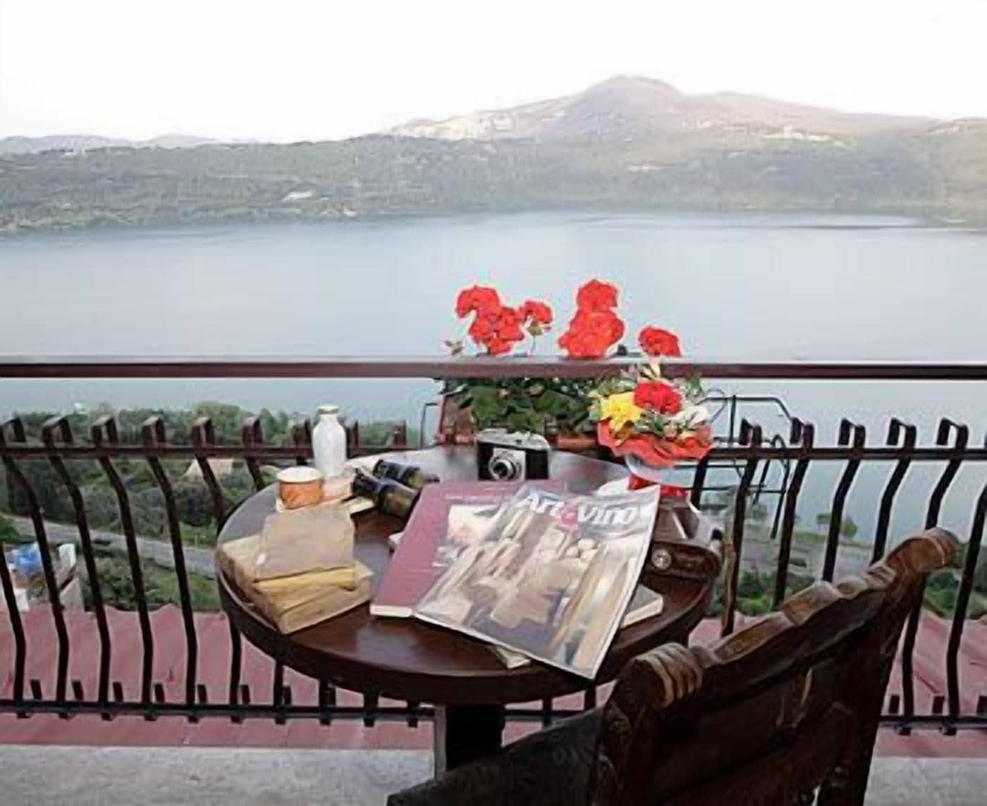 Una mesa con una revista, una cámara y un florero en un balcón con vista a un lago y montañas al fondo.