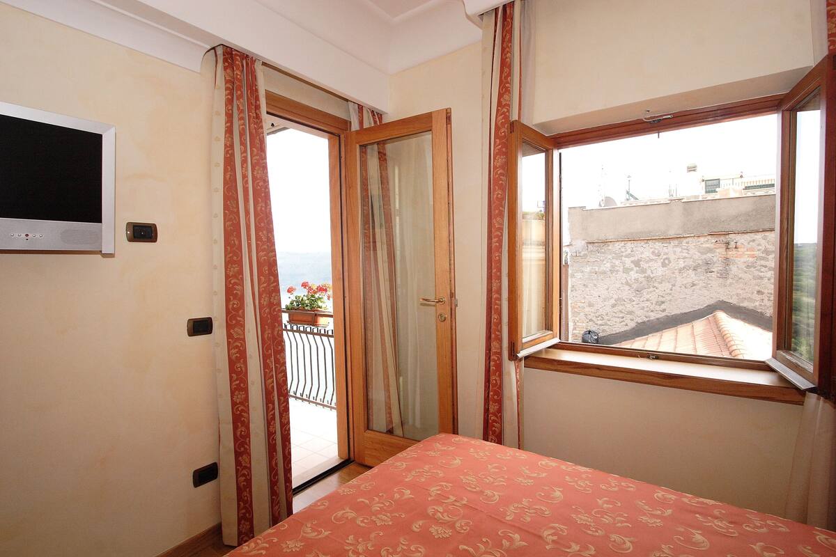 Un dormitorio con cama, televisor montado en la pared, ventana con vista a la ciudad y puerta que da a un balcón al aire libre.