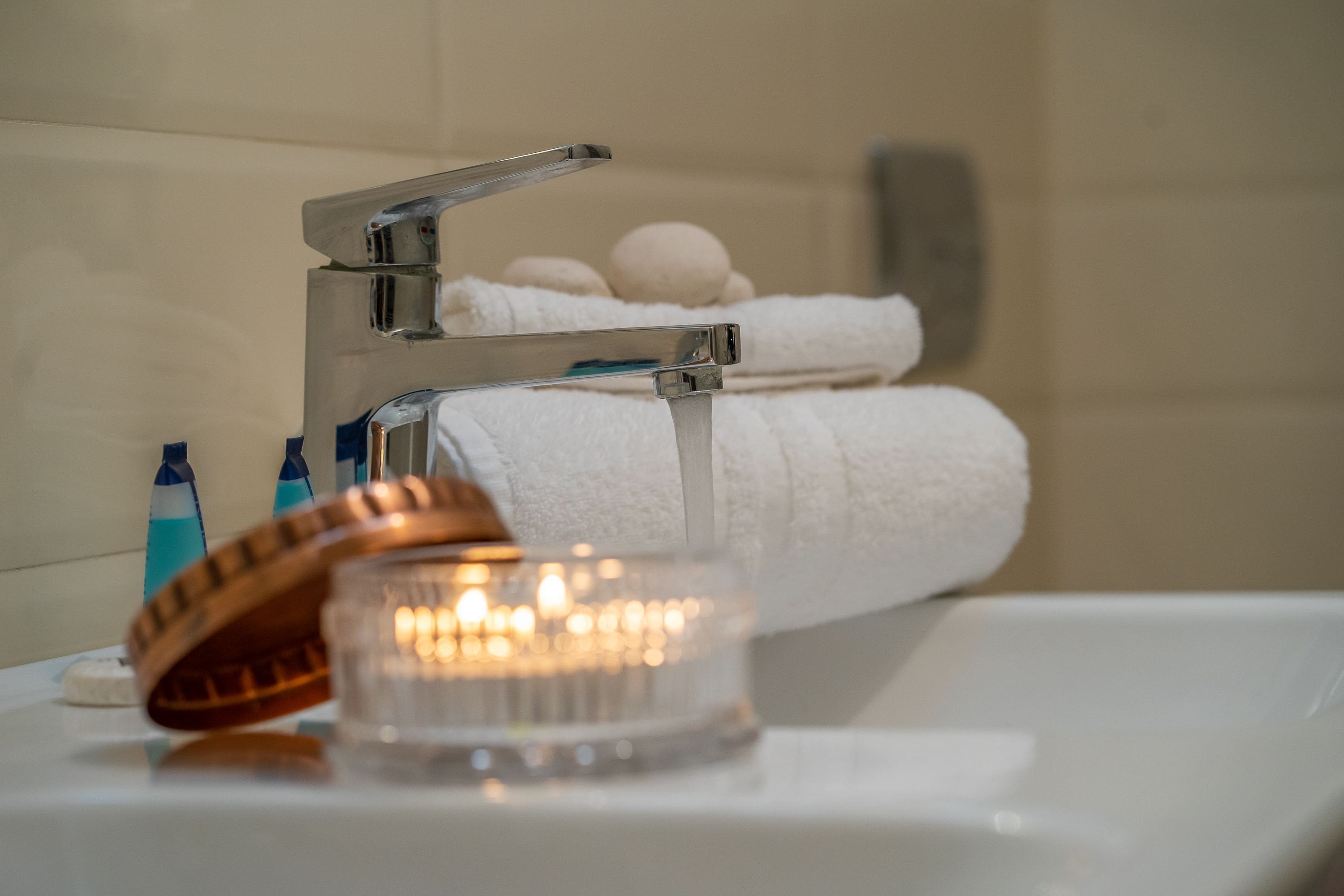 Un baño con un grifo moderno, una vela encendida y un perchero con toallas enrolladas.