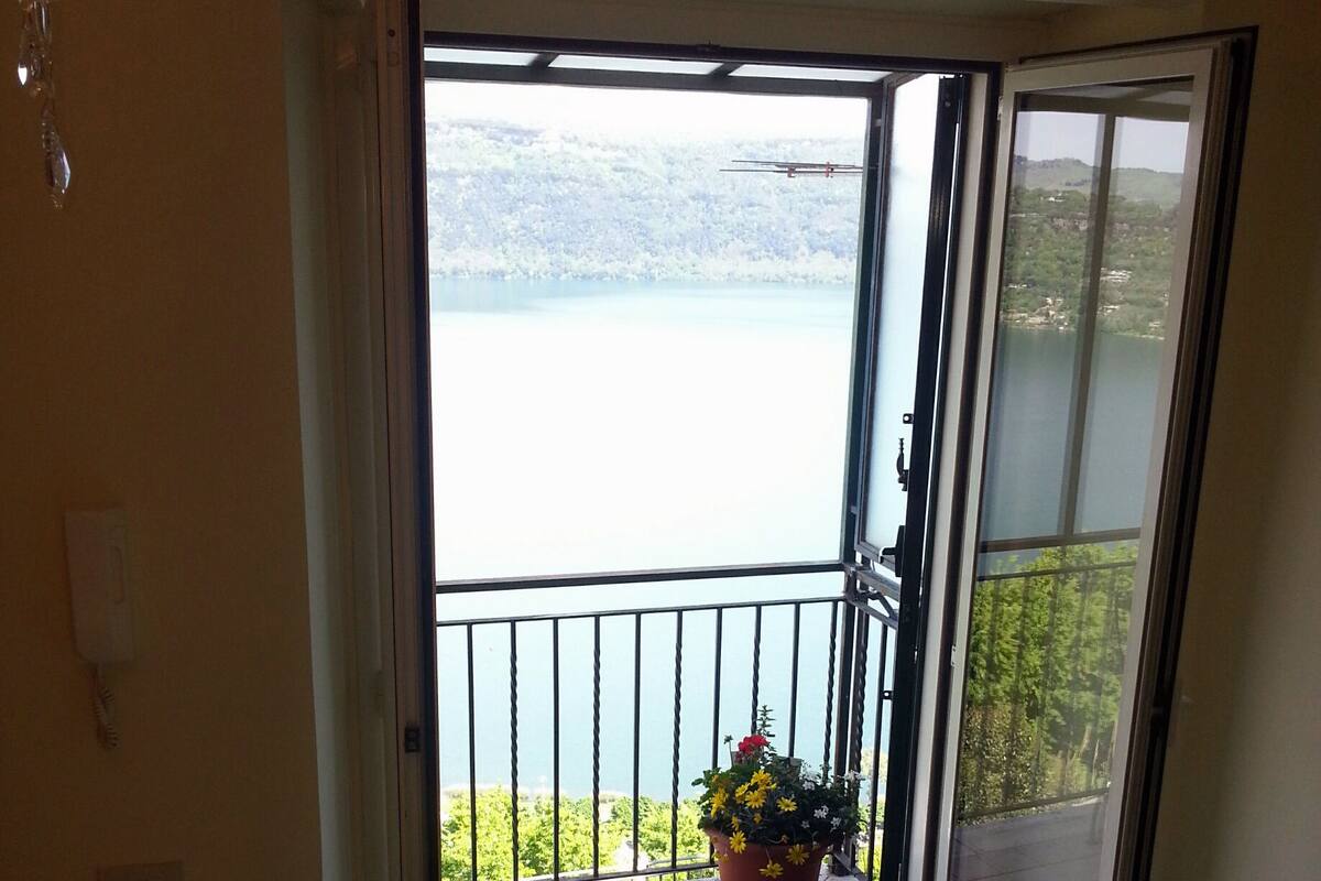 Una habitación con puerta corrediza de vidrio que da a un balcón con planta en maceta y vistas a un lago.