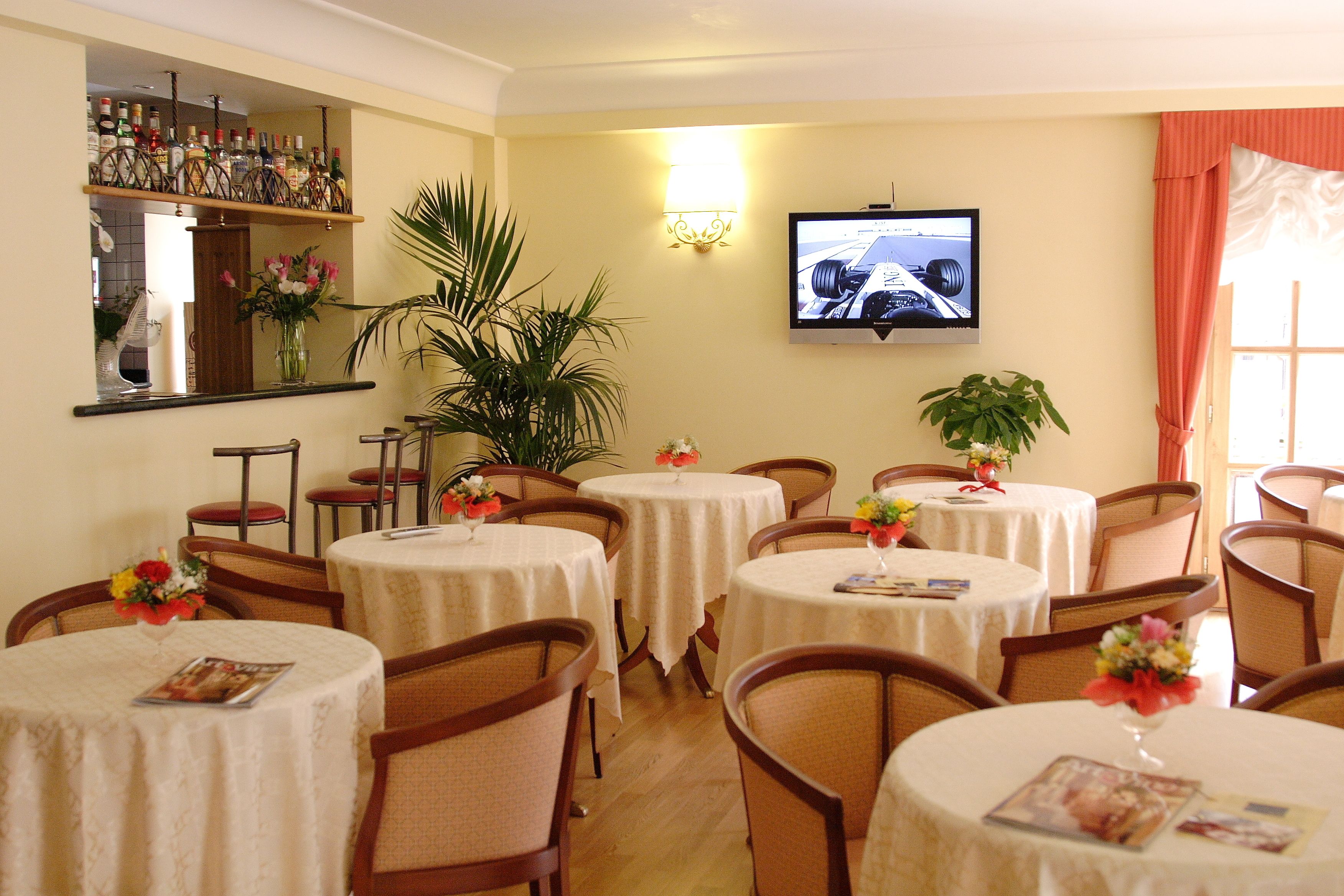 Un comedor con mesas redondas, sillas, un televisor montado en la pared y una zona de bar con botellas.