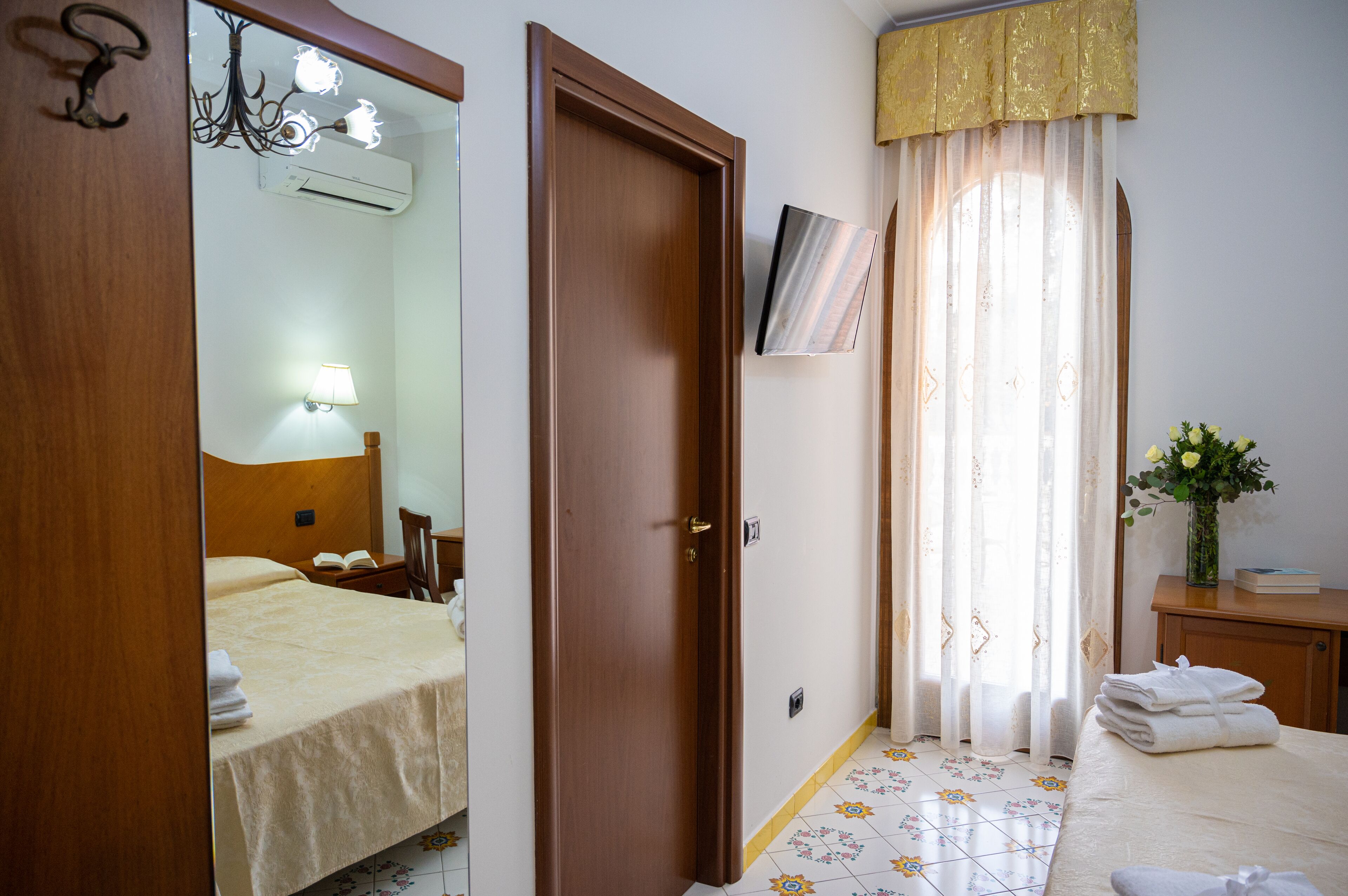 Una camera d'albergo con un letto, un comodino, uno specchio, una porta, una finestra con tende e un tavolino con un vaso di fiori.