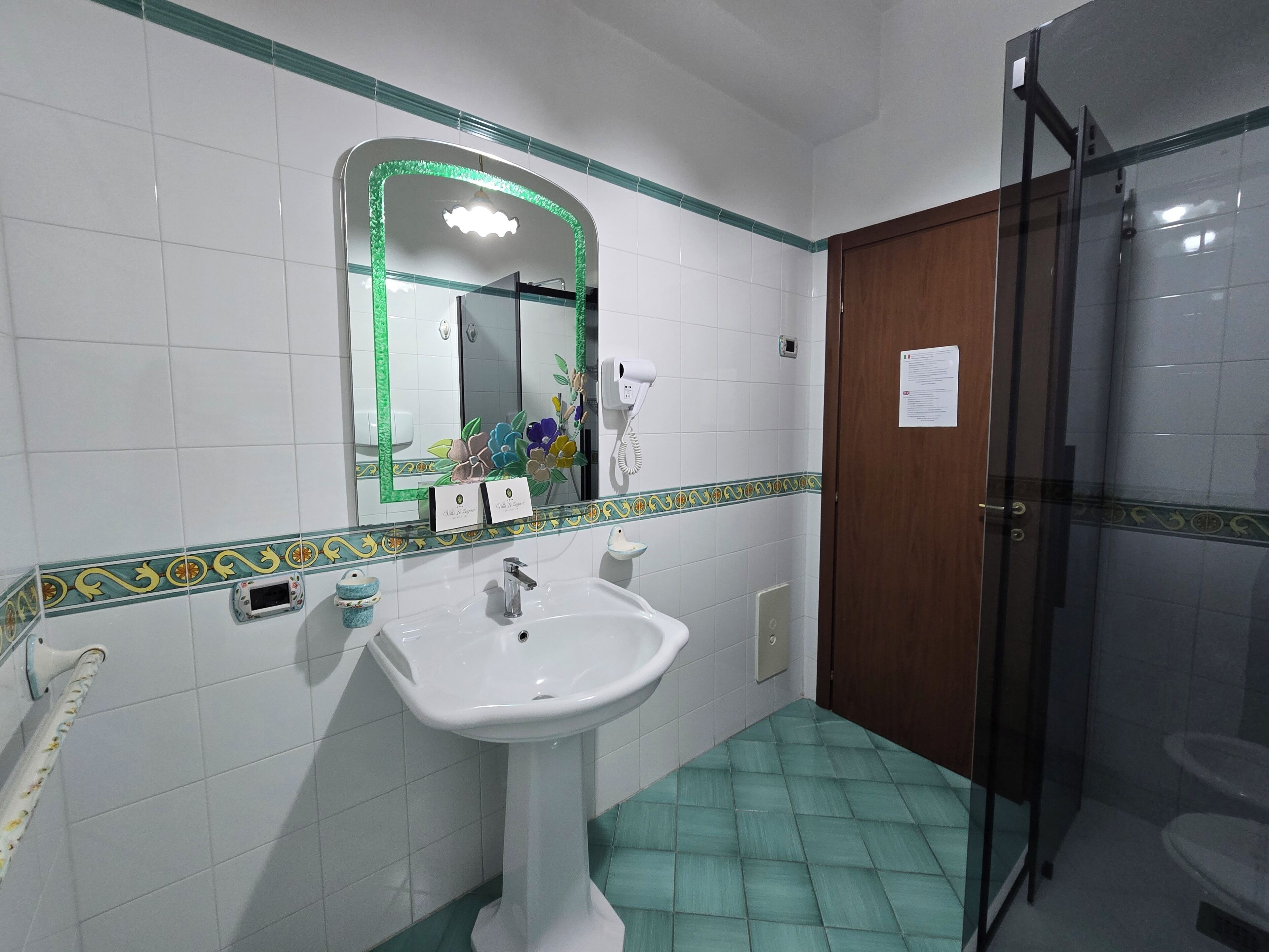 A bathroom with a white sink, a mirror with a green frame, a shower area, and a door with a sign.