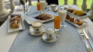 Une table dressée pour le petit-déjeuner, avec des viennoiseries, un assortiment de fruits et des boissons, donnant sur un jardin planté de palmiers.