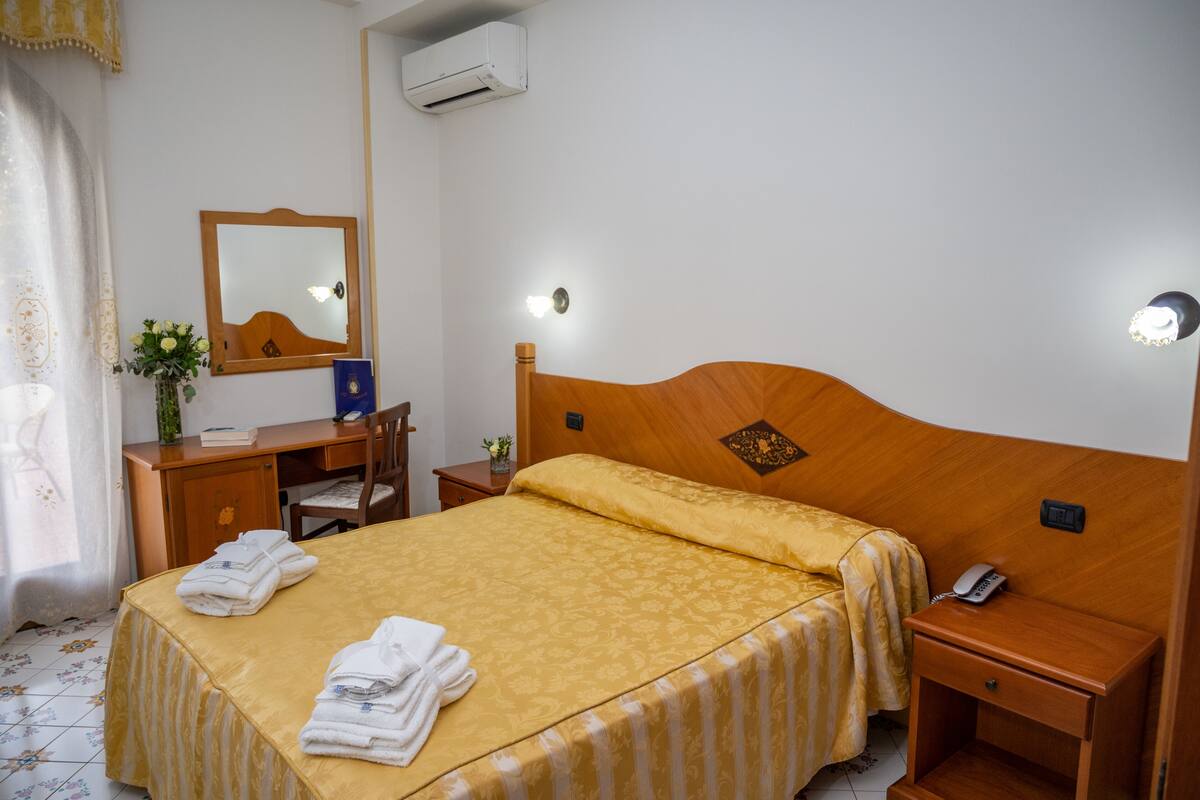 Een keurig opgeruimde hotelkamer met een bed, nachtkastje, een spiegel, een bureau met stoel en een kleine plant.