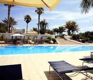 A pool area with lounge chairs and umbrellas, surrounded by palm trees and a building in the background.
