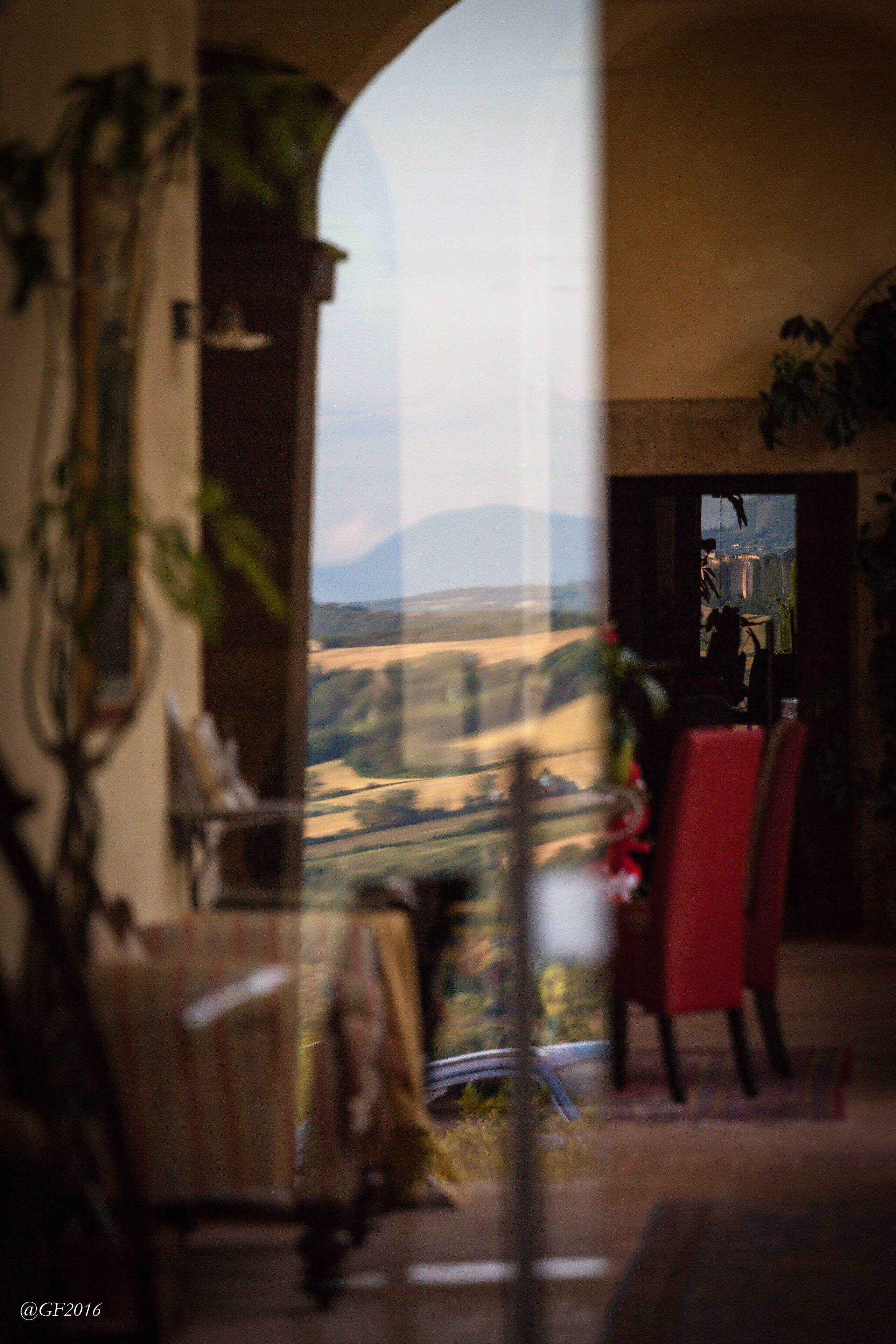 A room with a view of a landscape through a window. There is a table with chairs and a plant.