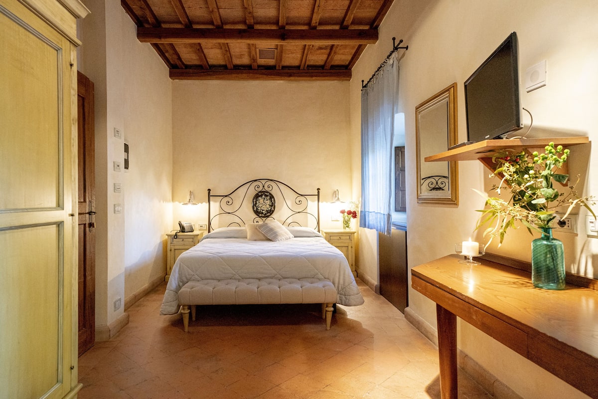 A bedroom with a bed, bedside tables, a nightstand, a TV on a shelf, and a vase with flowers.