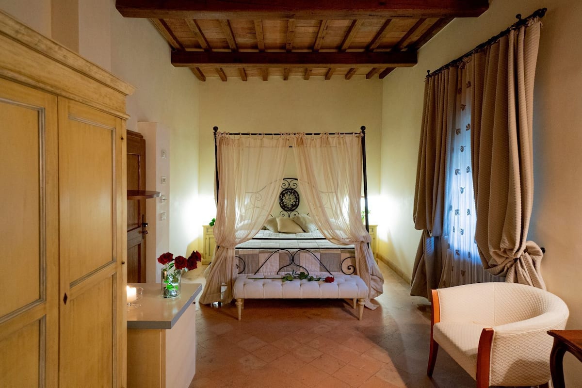 A bedroom with a four-poster bed, a wooden wardrobe, a bedside table with a vase of flowers, and a chair.