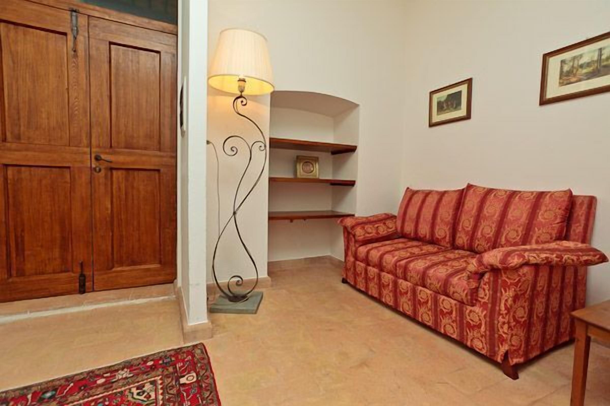 Ein Zimmer mit einem Holzschrank, einem roten, gemusterten Sofa, einer Stehlampe und einem kleinen Regal mit einem gerahmten Bild.