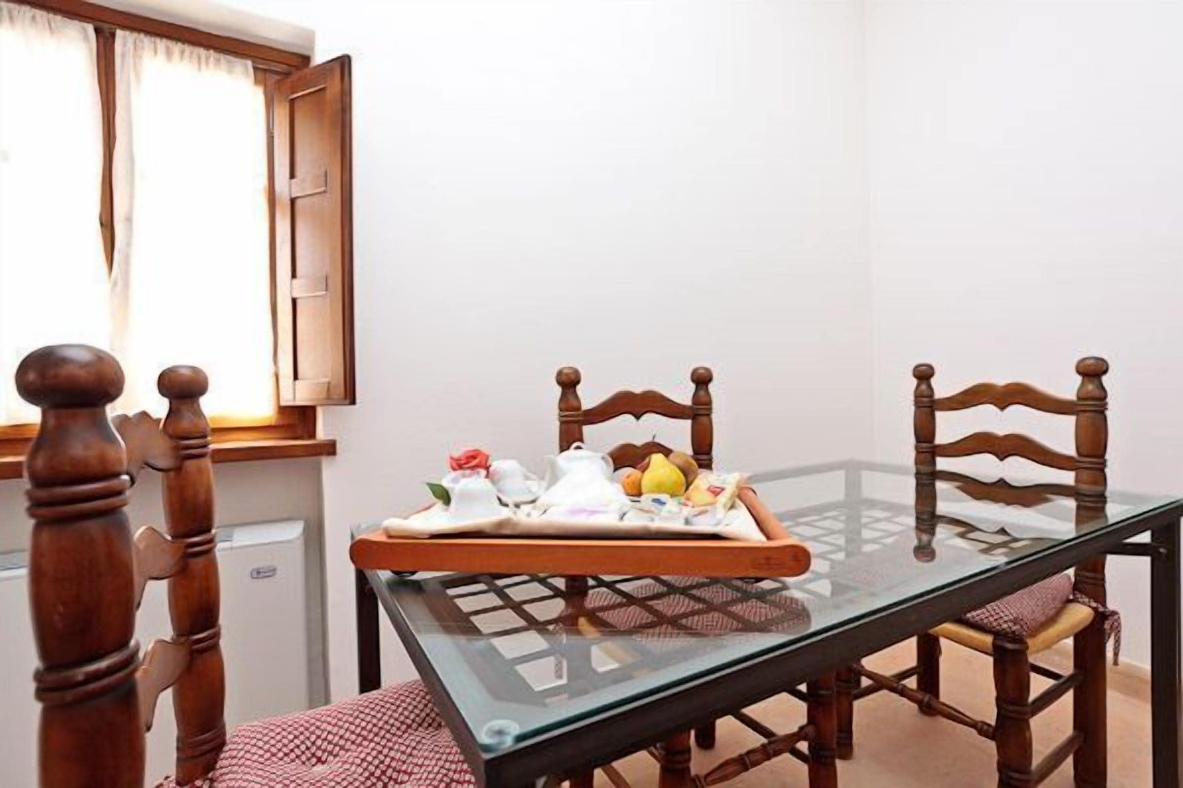 A dining area with a glass table, wooden chairs, and a tray with fruits and teapots.