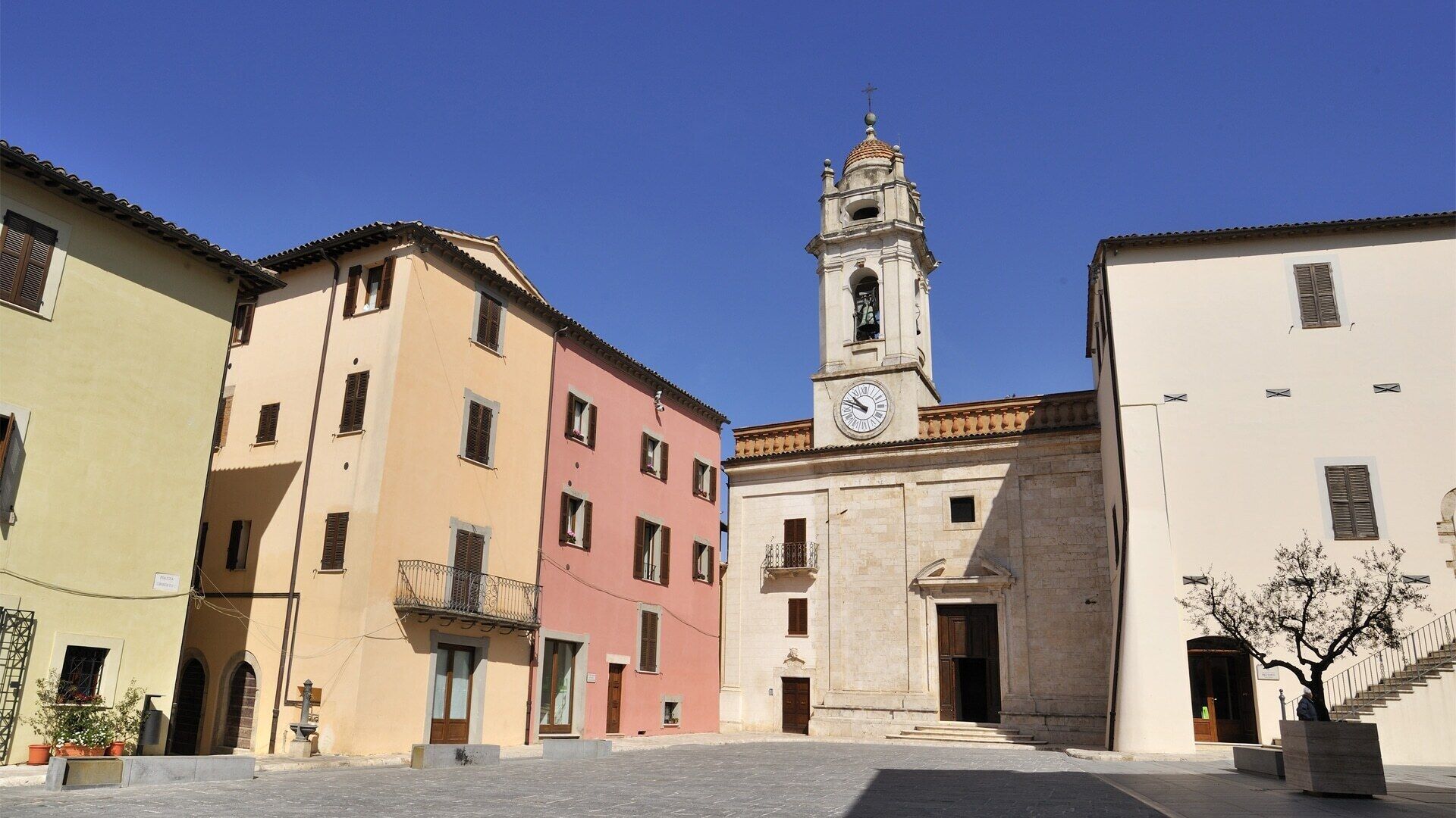 A square with buildings, a clock tower, and a tree.
