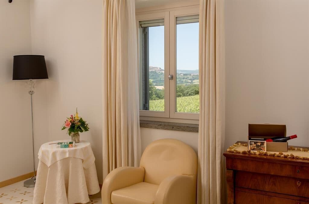 Une pièce avec vue sur un paysage verdoyant, visible à travers une fenêtre, comprenant une chaise, une table avec un vase de fleurs et une lampe.
