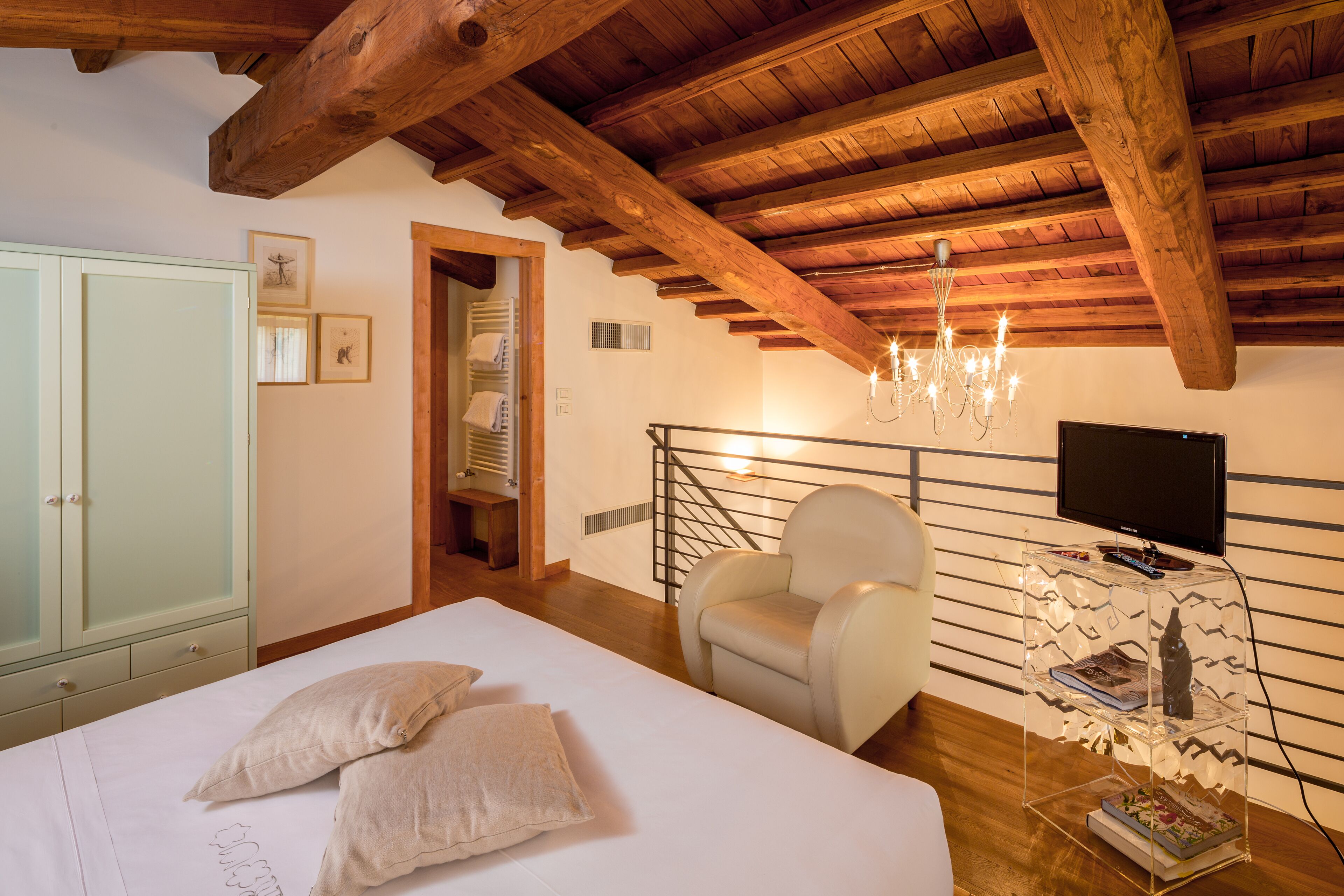 Een slaapkamer met een houten plafond, een bed met kussens, een stoel, een televisie op een glazen plank en een kledingkast.
