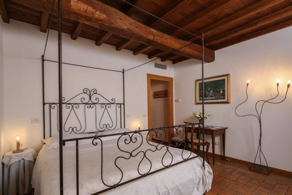A bedroom with a four-poster bed, a wooden ceiling, a framed cityscape painting, and a table with a vase of flowers.