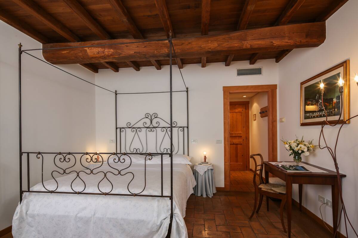 A bedroom with a four-poster bed, a wooden ceiling, a small table with a chair, and a framed picture on the wall.