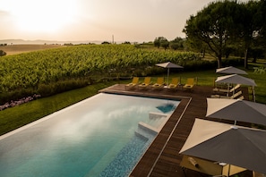 A rectangular infinity pool with a wooden deck, surrounded by lounge chairs and umbrellas, set against a backdrop of a vineyard and distant hills.