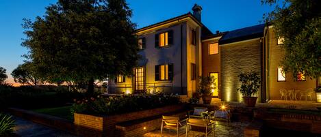 A large house with a stone patio, outdoor seating, and a well-lit garden at dusk.