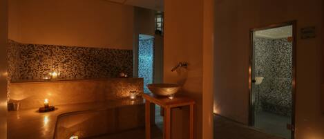 Zona de spa tenuemente iluminada con un muro de azulejos, una mesa de madera con un tazón y un banco.