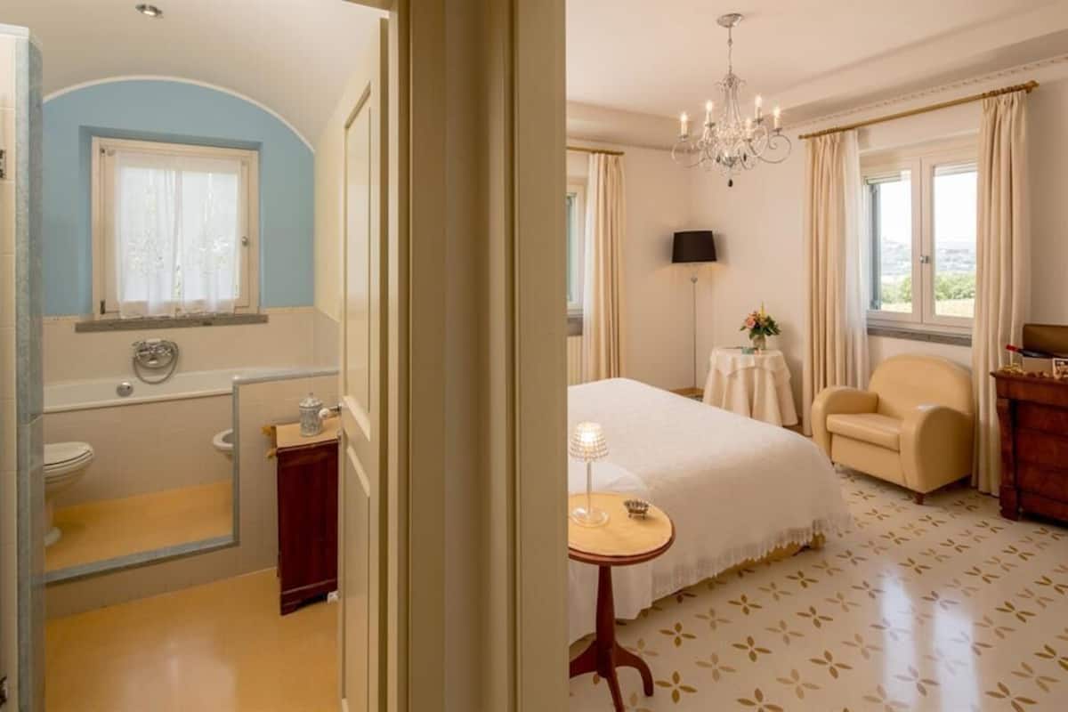A hotel room with a bathroom, a bed, a chair, a table, and a chandelier.