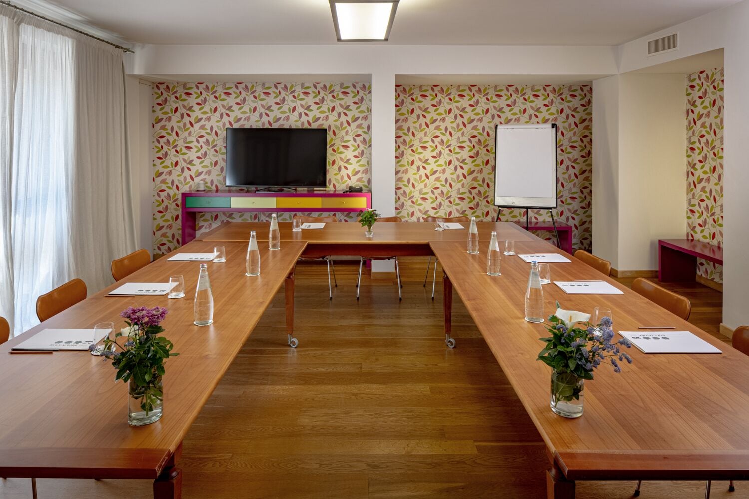 A conference room with a long wooden table, chairs, a TV, a whiteboard, and floral wallpaper.
