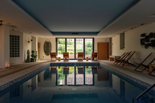 A pool area with a clear view of the outdoors, wooden lounge chairs, and a tiled floor.