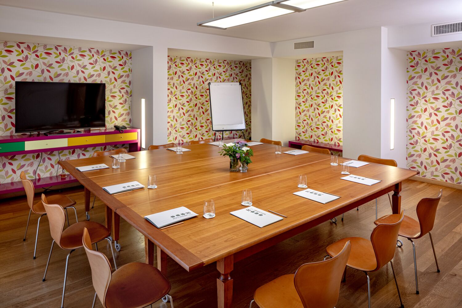 Une salle de conférence avec une longue table en bois, des chaises orange, un tableau blanc et un téléviseur fixé au mur.