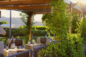 An outdoor dining area with tables set for two, covered by a wooden pergola, surrounded by lush greenery and a view of the cityscape.