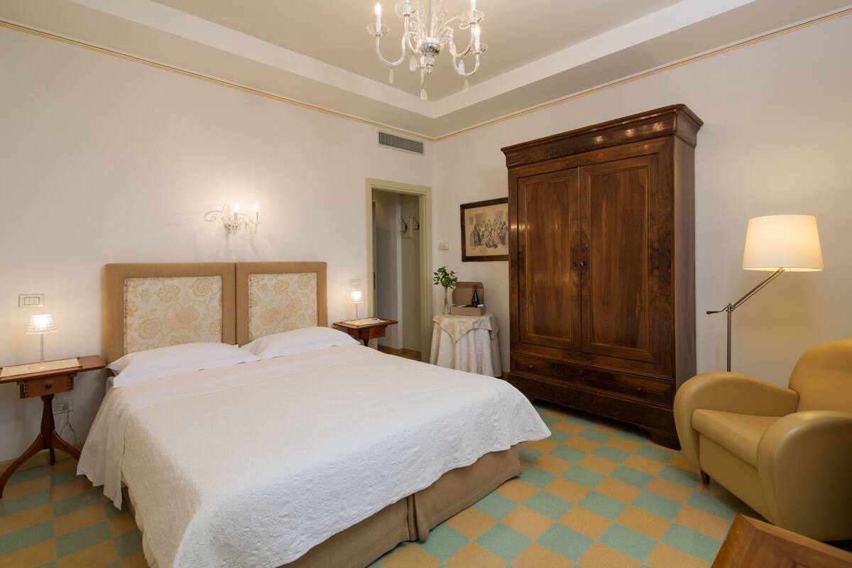 A hotel room with a large bed, a wooden wardrobe, a small table with a lamp, and a chair.