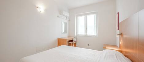 A bedroom with a bed, a desk, a chair, a window, and a wall-mounted air conditioner.