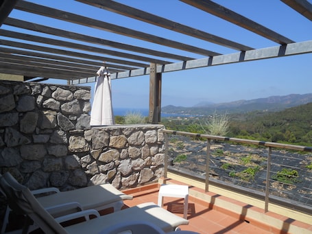 A balcony with stone walls, a pergola, and a view of the sea and hills.