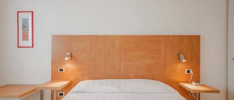 A modern bedroom with a wooden headboard, two bedside tables with lamps, a framed picture on the wall, and a small desk.