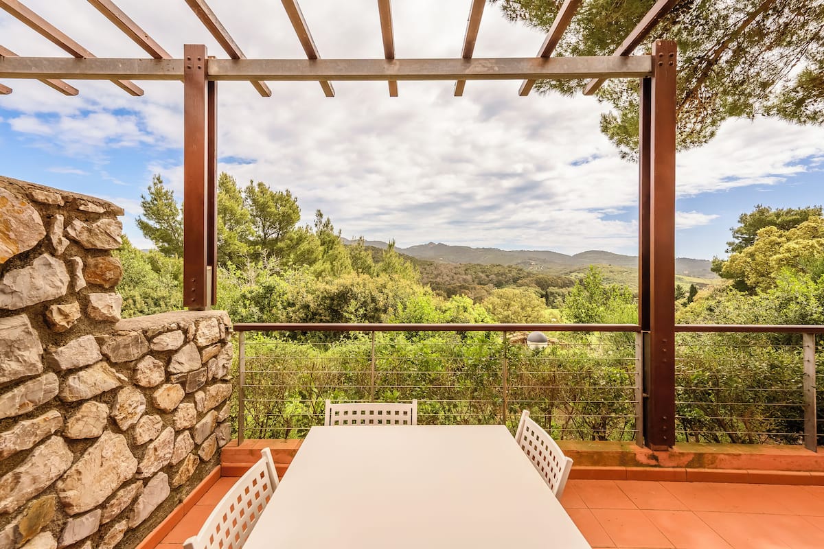 A balcony with a stone wall, white table, and chairs, overlooking a mountainous landscape.
