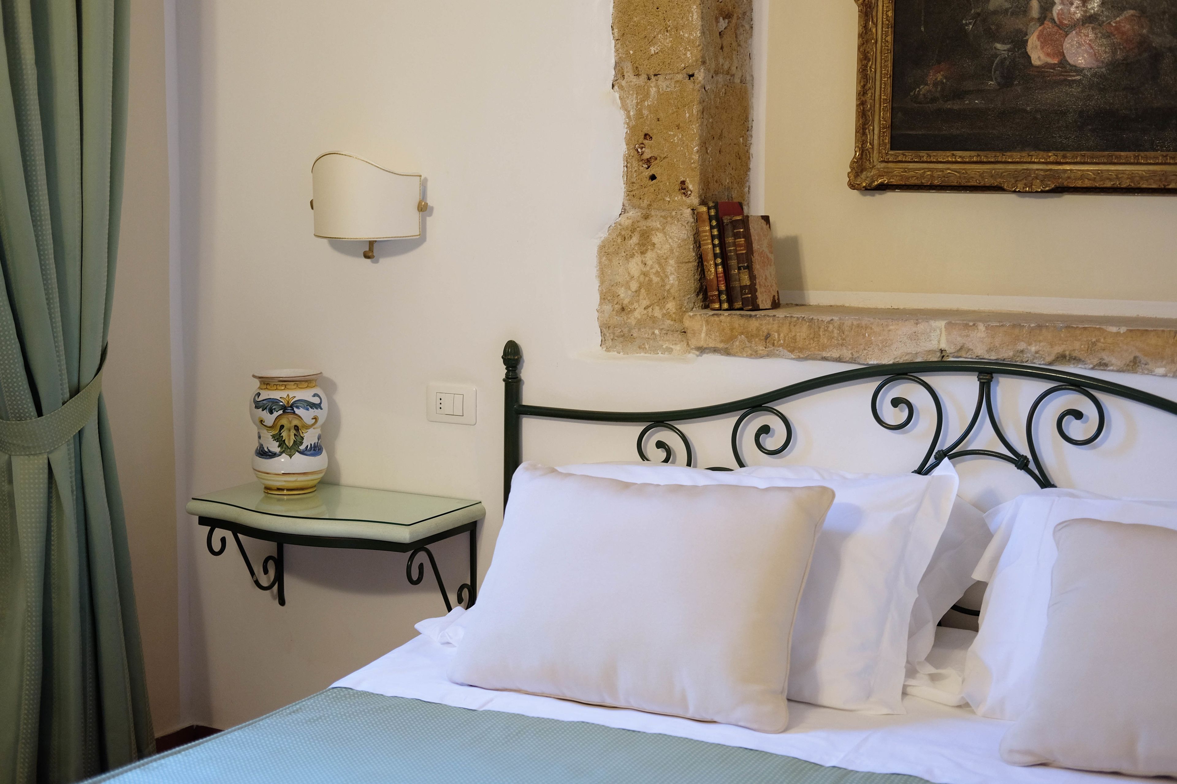 Una cama con sábanas blancas y varios almohadones, una mesita de noche con un florero, una lámpara de pared y una estantería con libros.