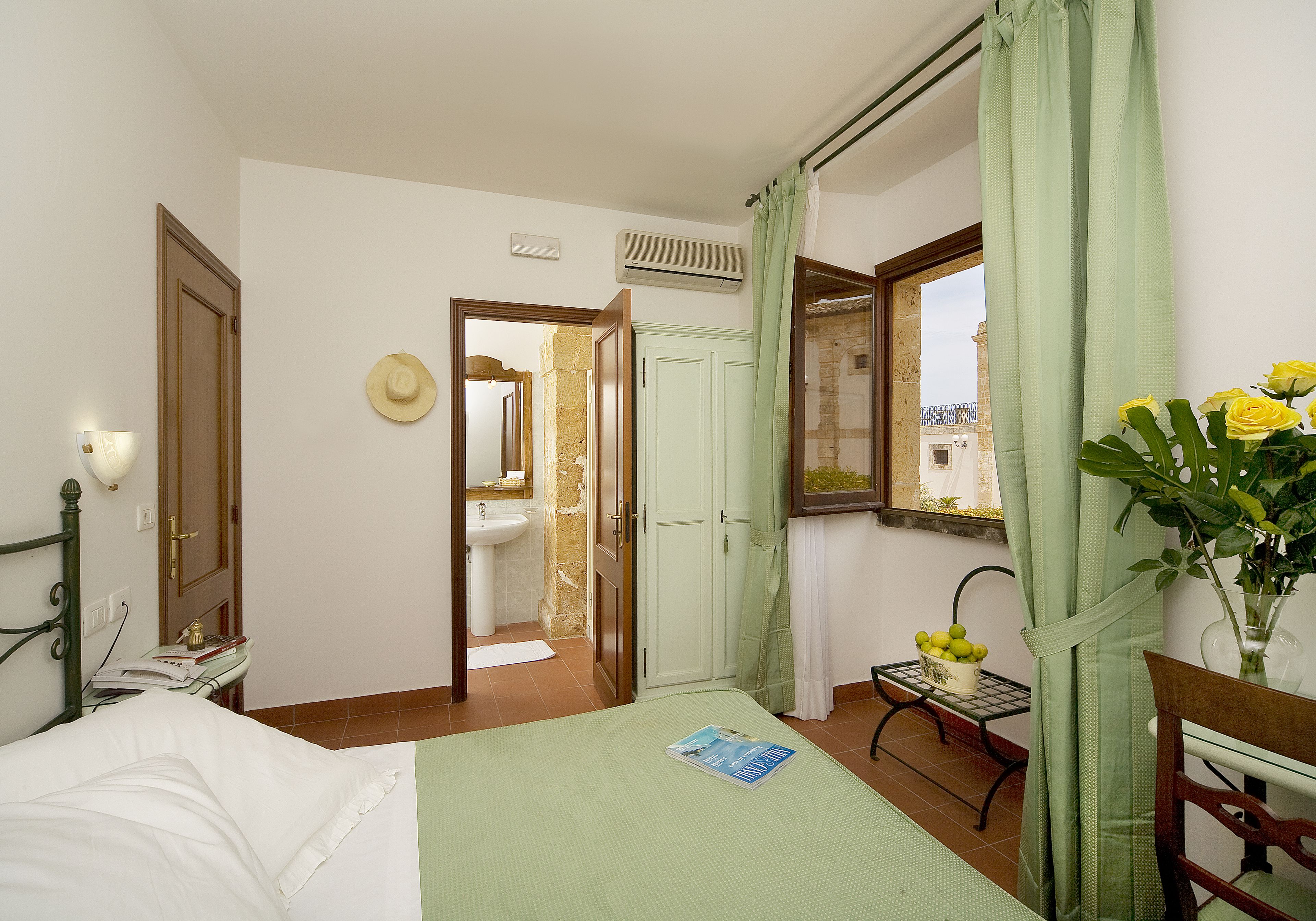 Un dormitorio con cama, una mesita de noche con un libro, una ventana con vistas y un baño visible a través de una puerta abierta.