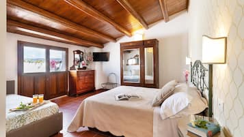 Un dormitorio con techo de madera, una cama con colcha color beige, una mesita de noche con un libro y flores, un aparador y un televisor montado en la pared.