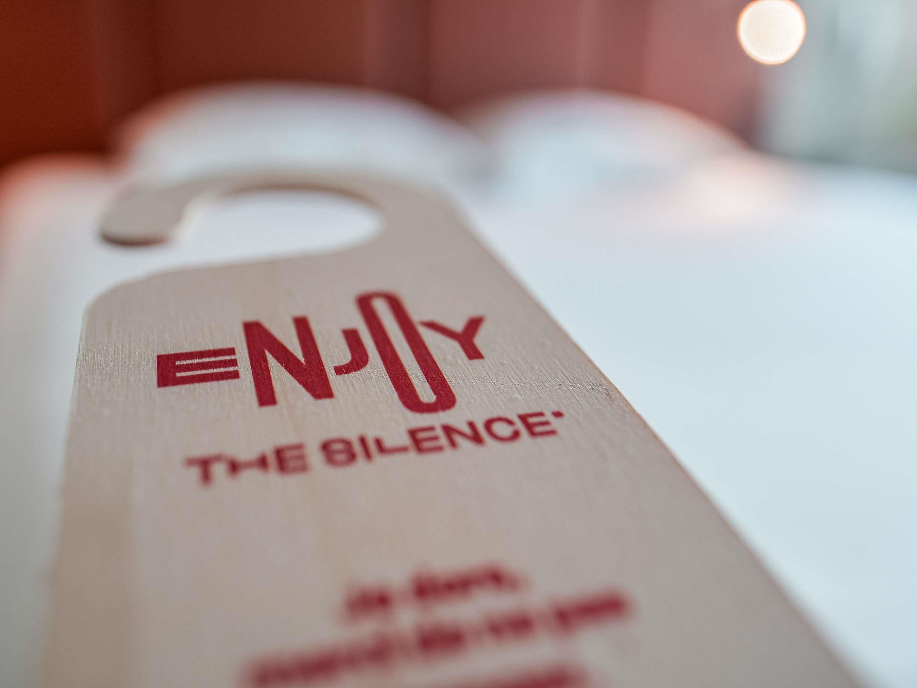 Primer plano de una etiqueta de madera con el texto 'ENJOY THE SILENCE' y una vista parcial de una habitación.