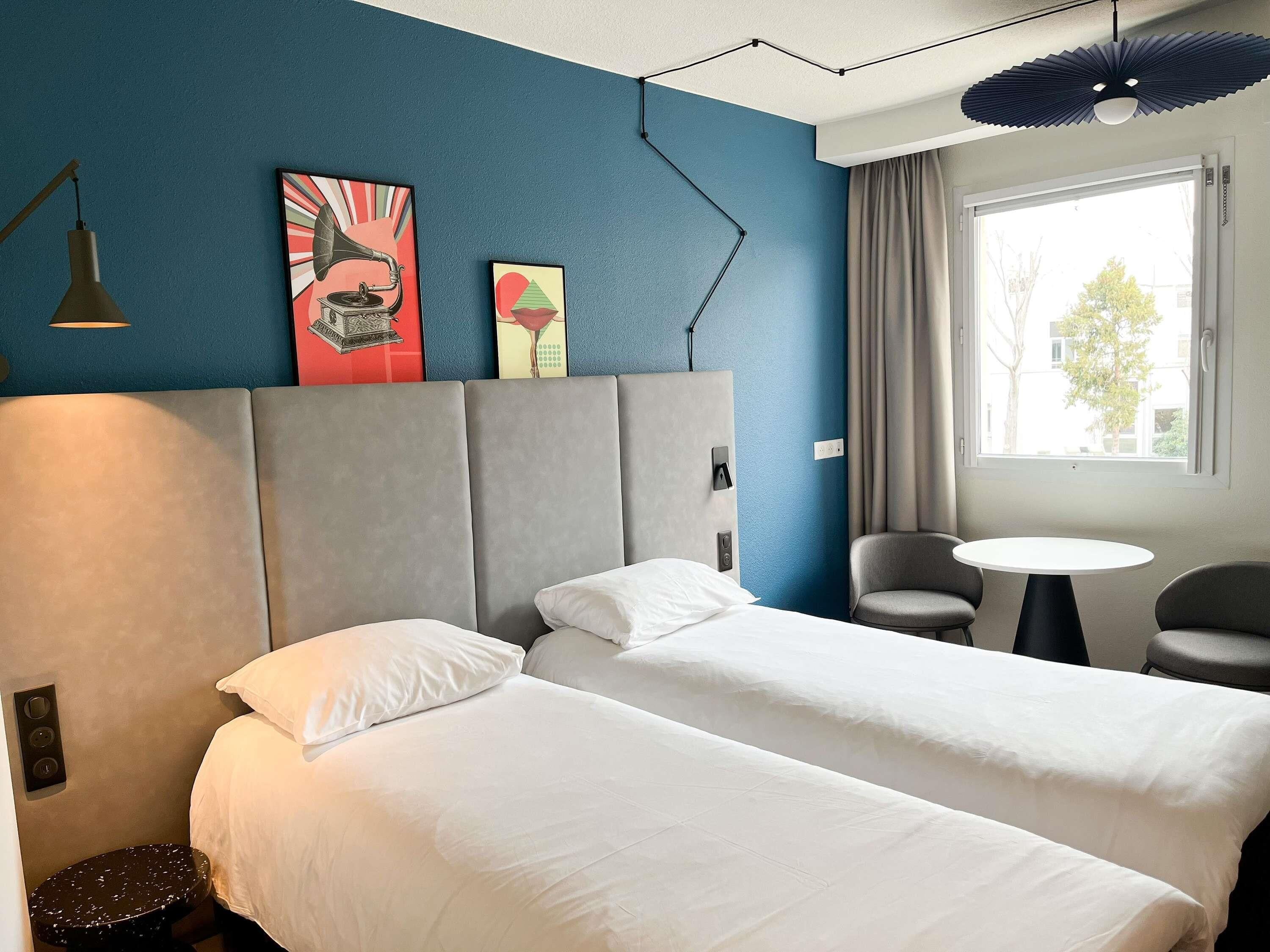 A hotel room with two beds, a desk, and a chair. There are two paintings on the wall, a window with curtains, and a ceiling fan.