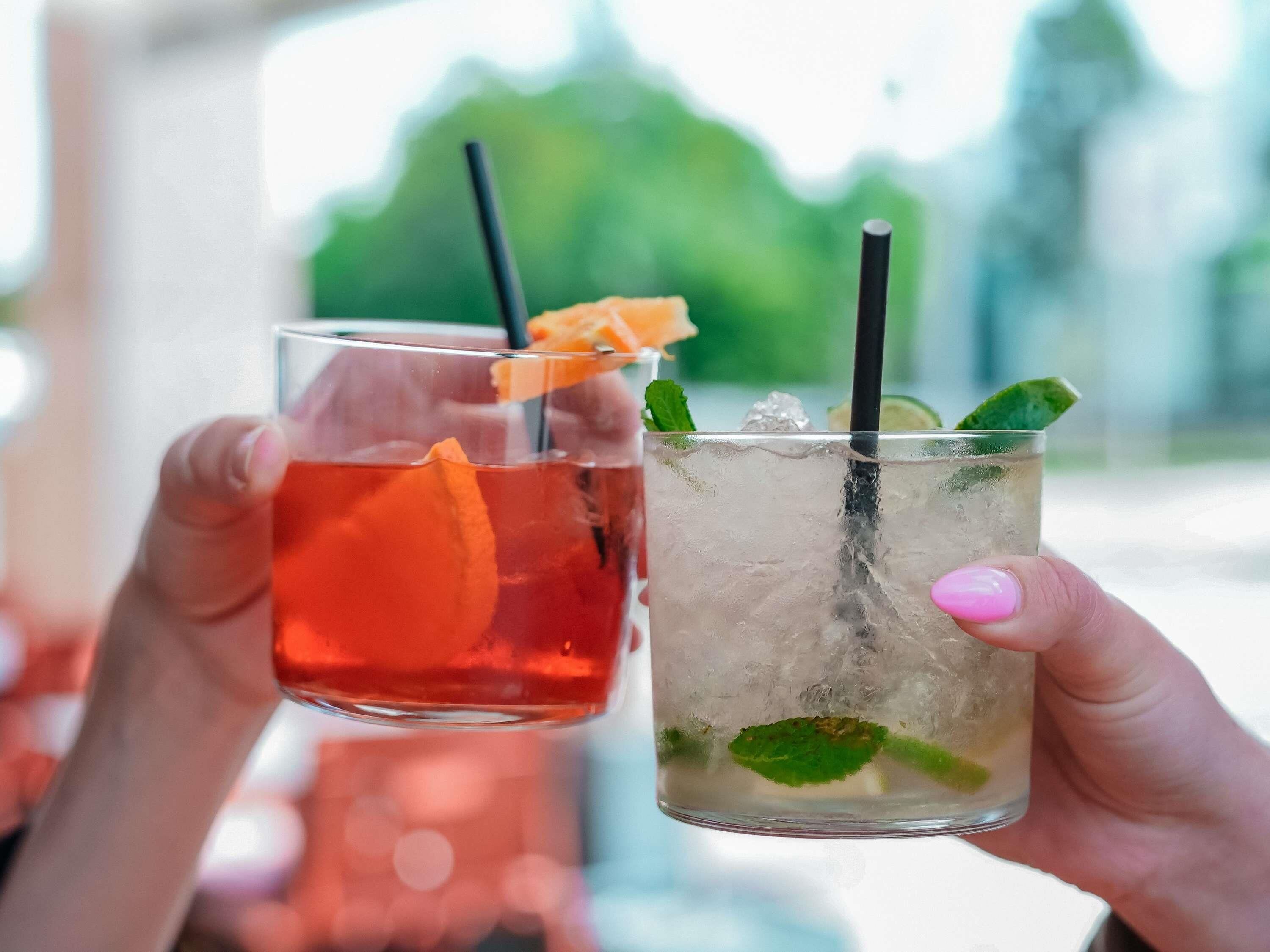 Two drinks with straws and garnishes, held by hands with pink nail polish.